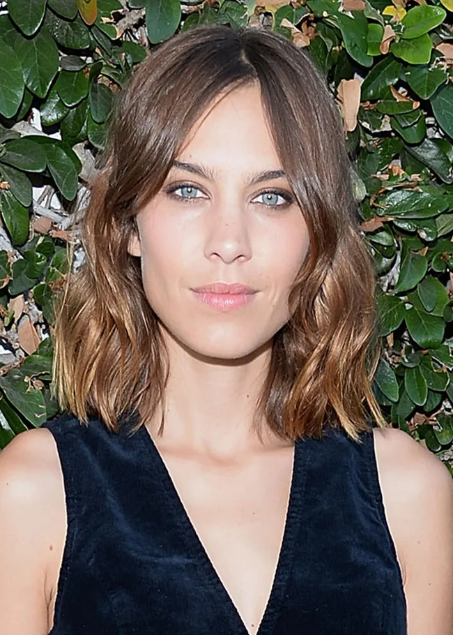 A woman with shoulder-length brown hair stands in front of green foliage, wearing a sleeveless dark blue top.