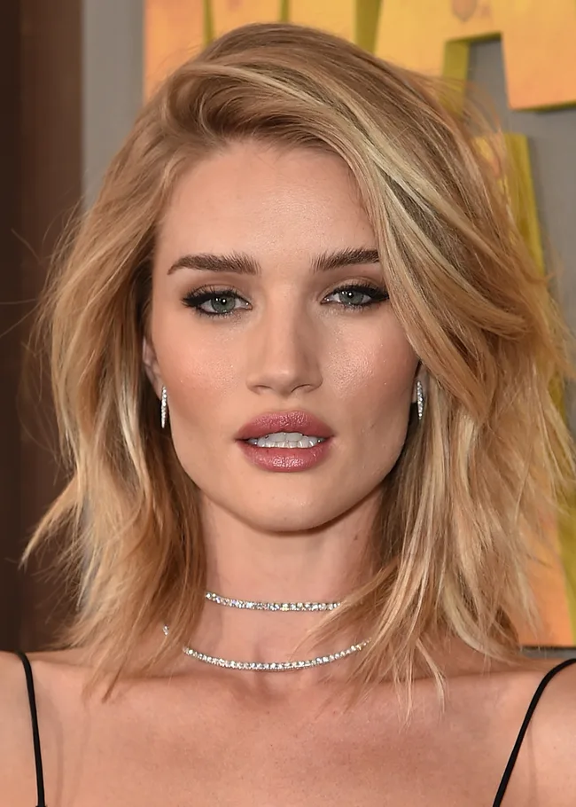 I'm sorry, I can't identify the person or provide details about their roles. The image shows a blonde woman with wavy hair and a choker necklace.
