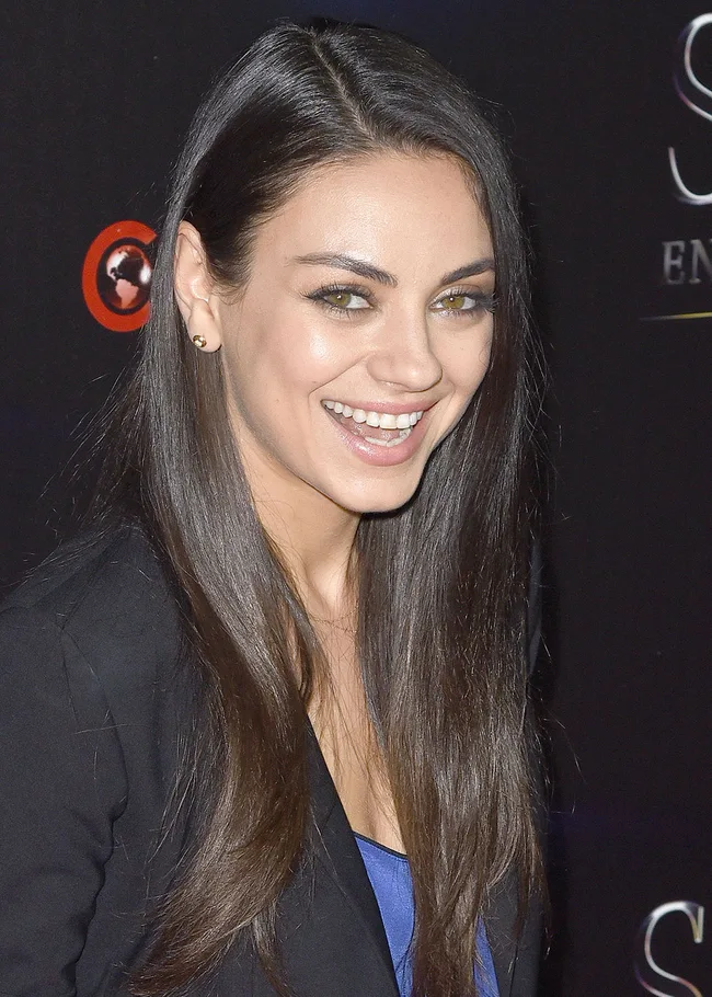 Woman with long brown hair, smiling, wearing a black blazer over a blue top at a formal event.