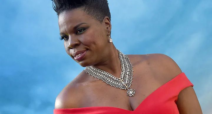 A person in a red off-shoulder dress poses in front of a blue background, wearing a layered diamond necklace.
