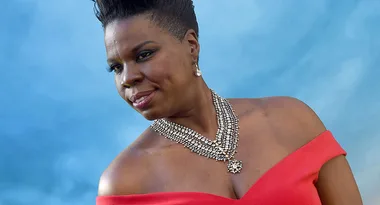 A person in a red off-shoulder dress poses in front of a blue background, wearing a layered diamond necklace.