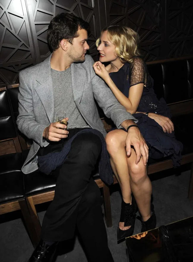 Man in gray blazer with drink and woman in black dress talking closely in a dimly lit room.