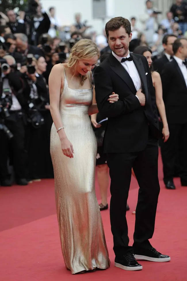 Couple walking on red carpet; woman in gold dress, man in black suit, surrounded by photographers.
