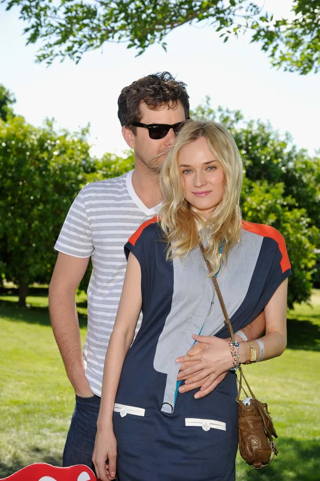 A man in sunglasses embraces a smiling woman outdoors with trees and grass in the background.