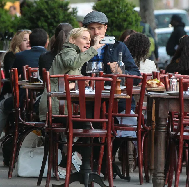 Couple at a cafe taking a selfie with a smartphone, surrounded by red chairs and tables, enjoying outdoor dining.