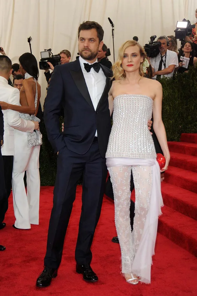 Couple on red carpet; man in black tuxedo, woman in strapless white beaded gown with sheer pants, surrounded by press.