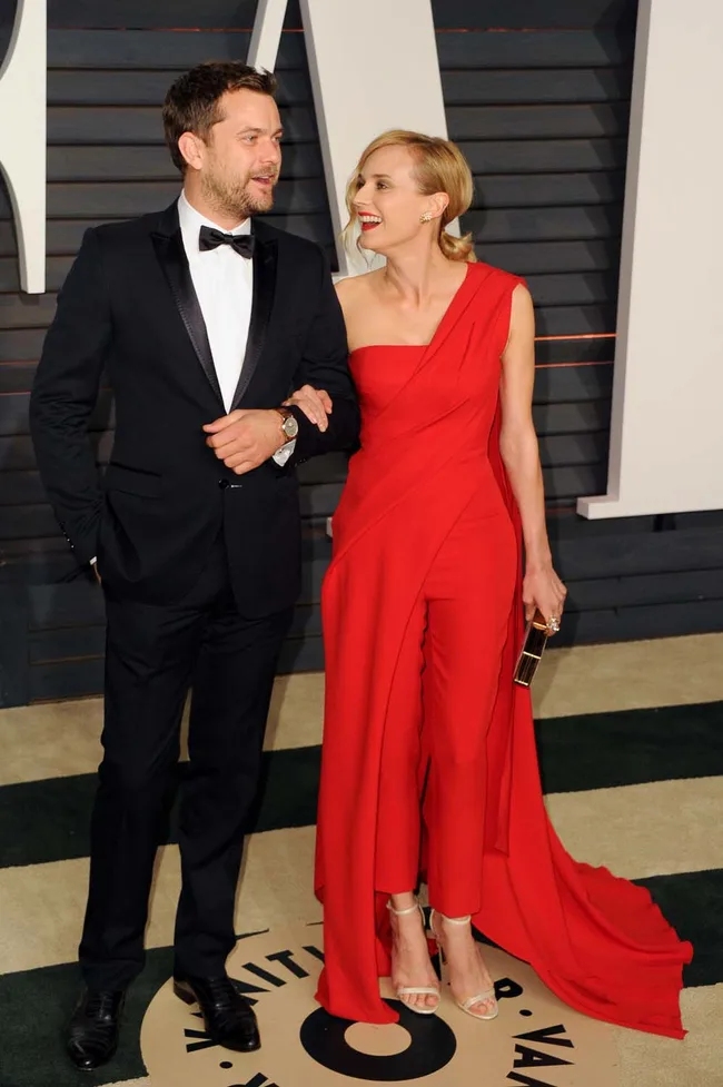 A couple in formal attire on a red carpet; man in a black tuxedo, woman in a red dress with a flowing train.