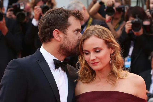 Man whispering to woman on red carpet, surrounded by photographers.