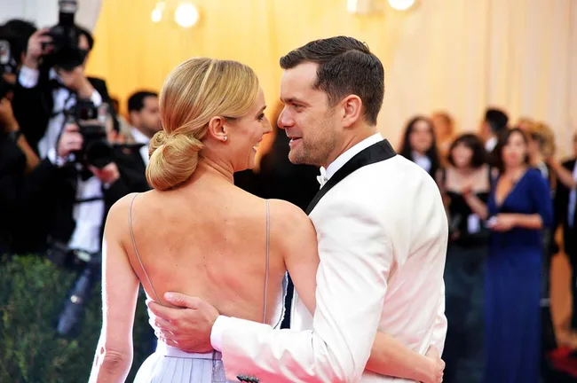 A couple in formal attire embraces on the red carpet, surrounded by photographers.
