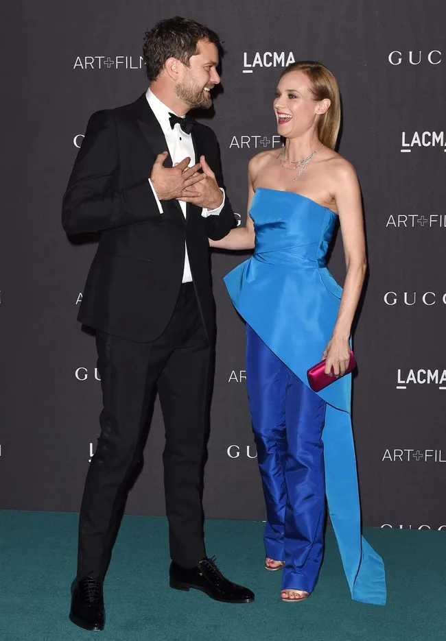 A man in a black suit and a woman in a blue gown converse on the red carpet at an event.