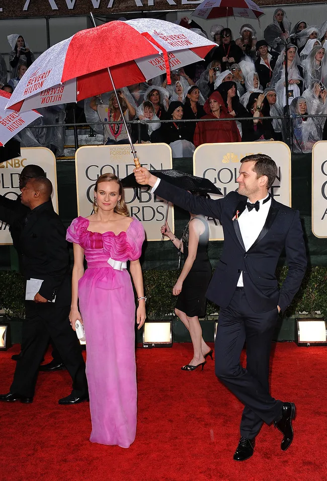 Man holds umbrella for woman in a pink dress on a rainy red carpet at the Golden Globe Awards.