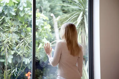 Woman with long blonde hair gazing out a window into a lush green garden.