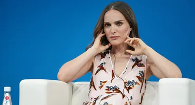 A woman in a floral dress sits on a white chair against a blue background, adjusting her earpiece.