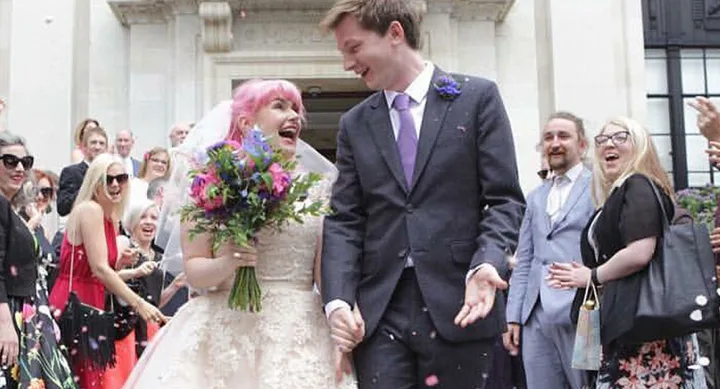 Bride with pink hair and groom laughing joyfully, holding hands and bouquet, surrounded by cheering guests at a wedding.