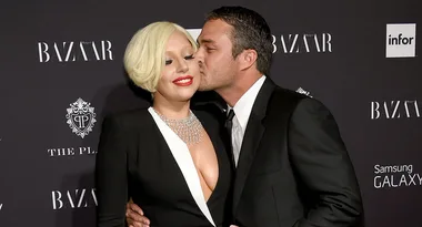 A man in a suit kisses a smiling woman in a low-cut dress on a Harper's Bazaar event red carpet.
