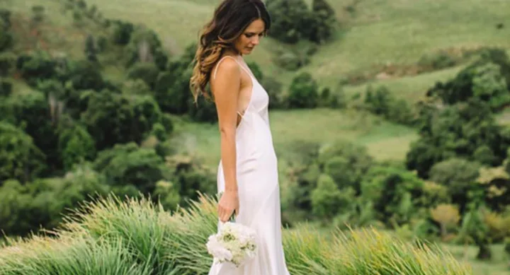 Bride in a white gown holding flowers, standing on grassy hillside with scenic green landscape in the background.