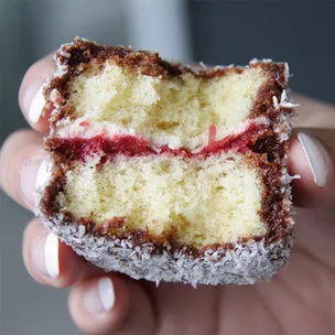 Close-up of a hand holding a sliced lamington cake with chocolate, coconut, and a red jam filling.