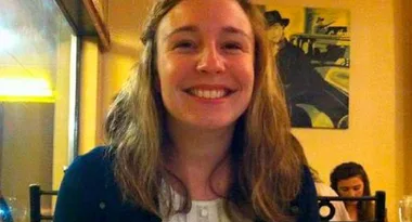 A woman with long hair smiling indoors with a portrait in the background.