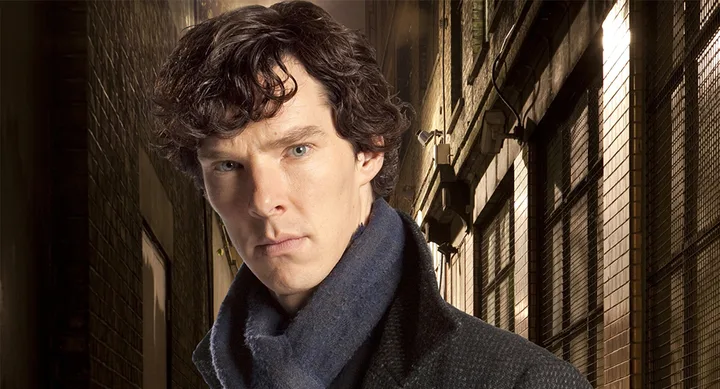 Man in a dark coat and scarf stands in a dimly lit alleyway, looking serious and focused.