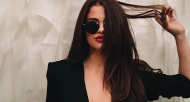 A woman wearing sunglasses, in a black outfit, playfully holds her hair against a textured cream-colored backdrop.