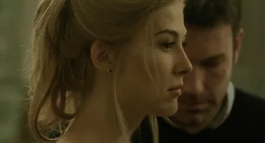 A woman in profile faces left while a man stands behind her, both in a dimly lit room.