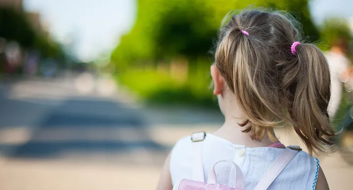 Young girl with blonde pigtails and a pink backpack walking outdoors on a sunny day.