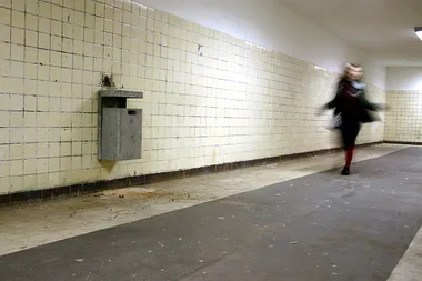 Blurred person running through a dimly lit tiled hallway, trash bin on the left wall.