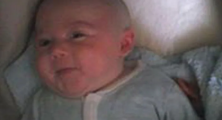 Smiling baby lying down in light-colored clothing, partially covered with a blanket.