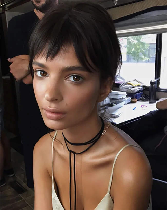 A woman with dark hair and bangs, wearing a thin strap top and a black choker, sits indoors near a table with items.