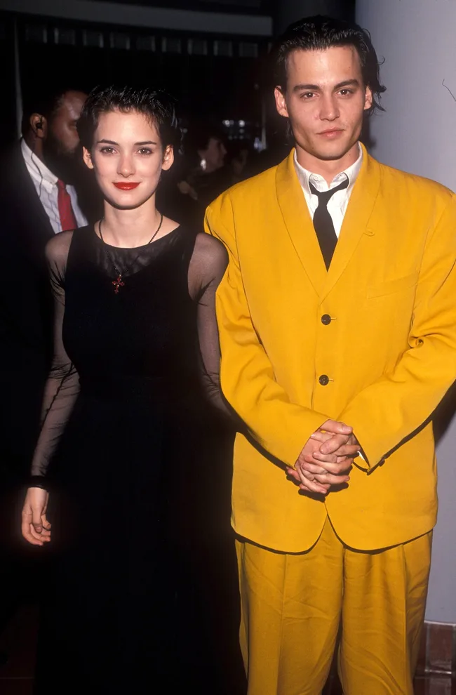 A person in a black dress stands with another in a yellow suit at an event, both with short dark hair.