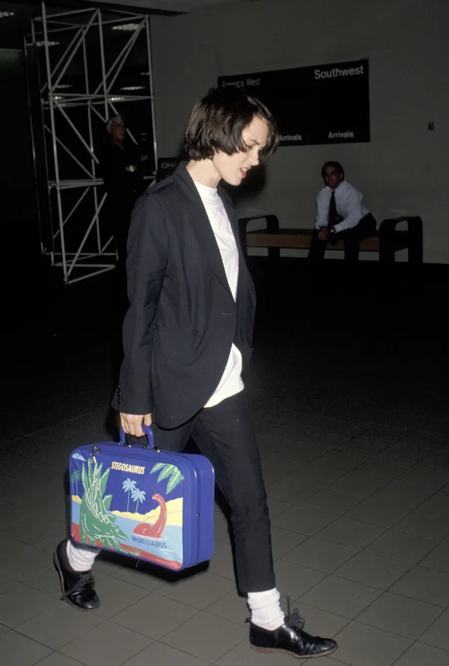 Person in black suit walking with a colorful dinosaur-themed suitcase at an airport.
