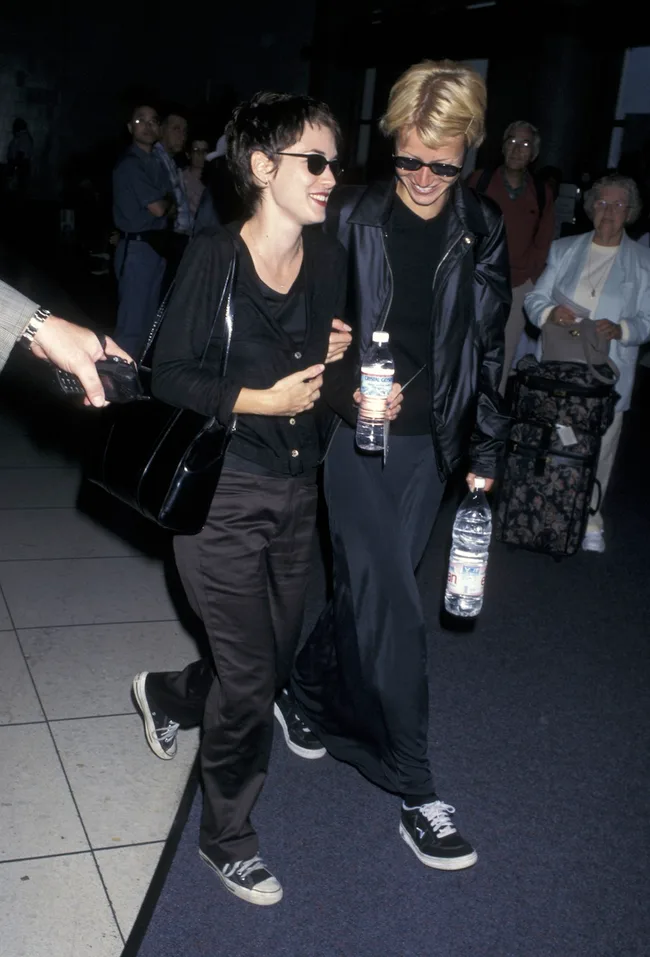 Two people wearing sunglasses and casual outfits walking together, smiling, and holding water bottles indoors.