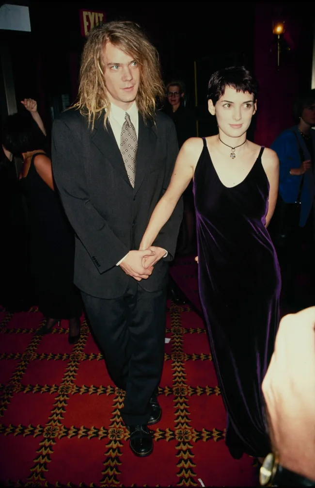 A couple holds hands, man in suit, woman in dark dress, walking on a patterned carpet at an event.