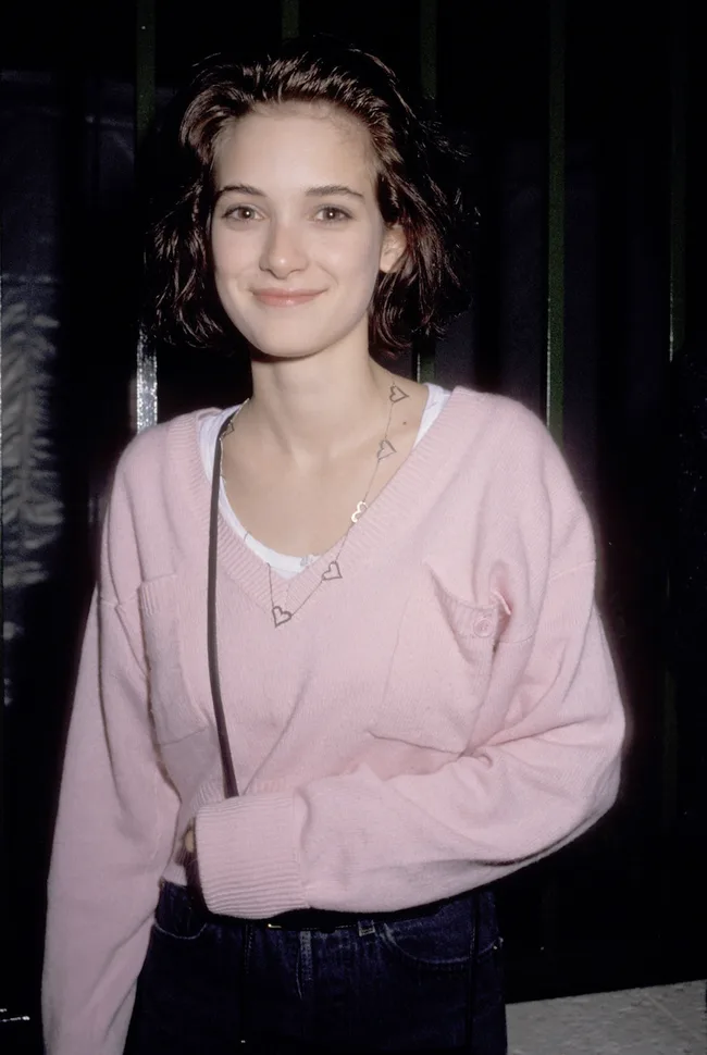 A person wearing a pink sweater and heart necklace smiles at the camera.