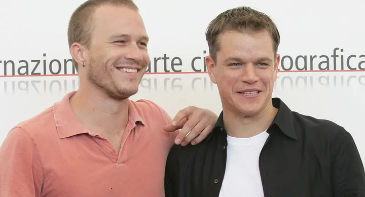Two smiling men stand together, one in a pink shirt and one in a black shirt, in front of a festival backdrop.