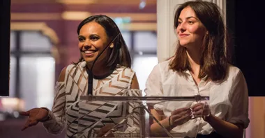Two women stand behind a podium speaking, one in a patterned dress, the other in a white shirt, smiling at an event.