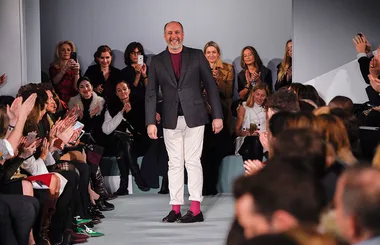 A man in a blazer and pink socks walks down a fashion runway as the seated audience applauds.