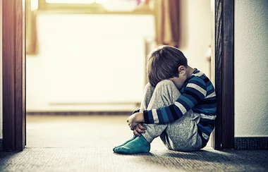 Child sitting on the floor in a hallway, hugging knees, wearing striped shirt and socks, looking down.