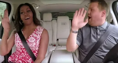 Two people in a car, smiling and raising hands as if waving or dancing, wearing seatbelts.