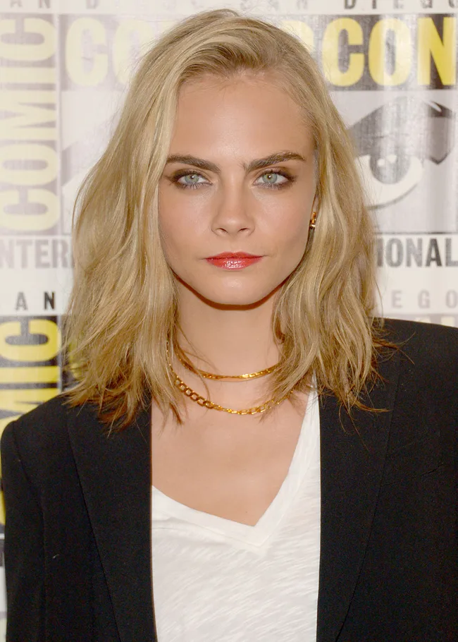 Woman with blonde hair, wearing a black blazer and gold necklaces, at Comic-Con event.