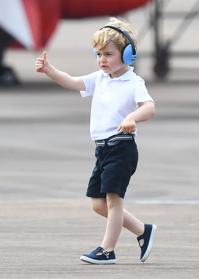 Young child in a white shirt, blue shorts, and earmuffs, giving a thumbs up while walking outdoors.