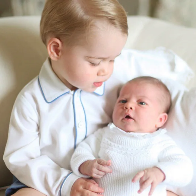 A young boy in a white shirt holds a baby in white clothes, both sitting on a white couch.