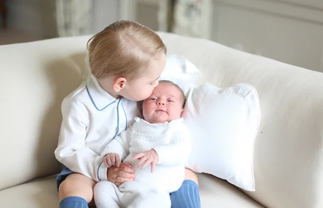 Toddler kisses baby on a white couch, both wearing white outfits; baby rests head on a pillow.