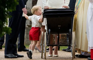 Toddler in red shorts holds a man's hand and stands beside a vintage black pram with adults around.