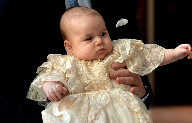 Baby in an ornate lace gown is held, showing detailed clothing and a calm expression.
