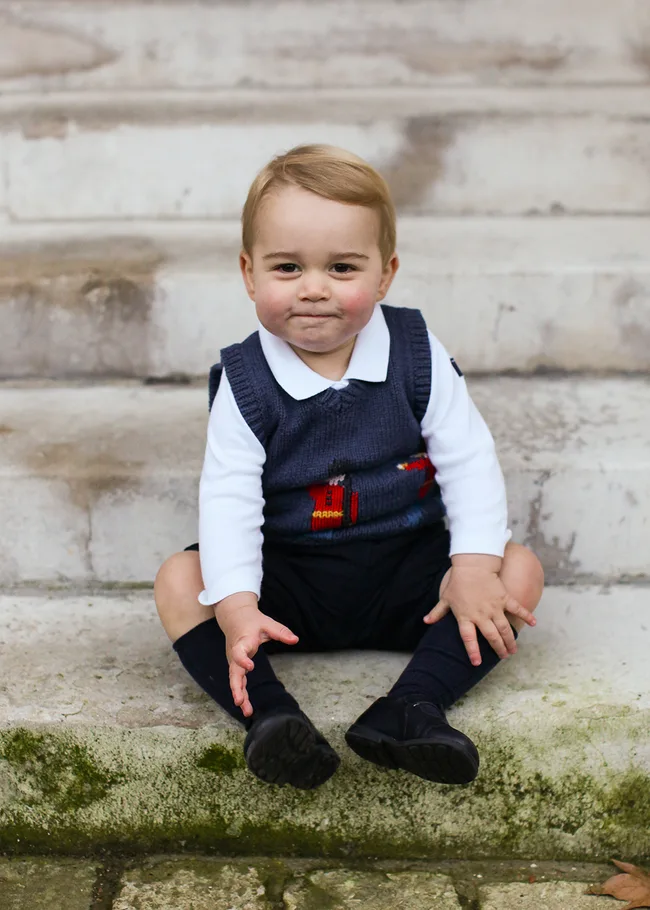 A young child sits on outdoor steps in a knit sweater, white shirt, and black shoes, with a slight smile.