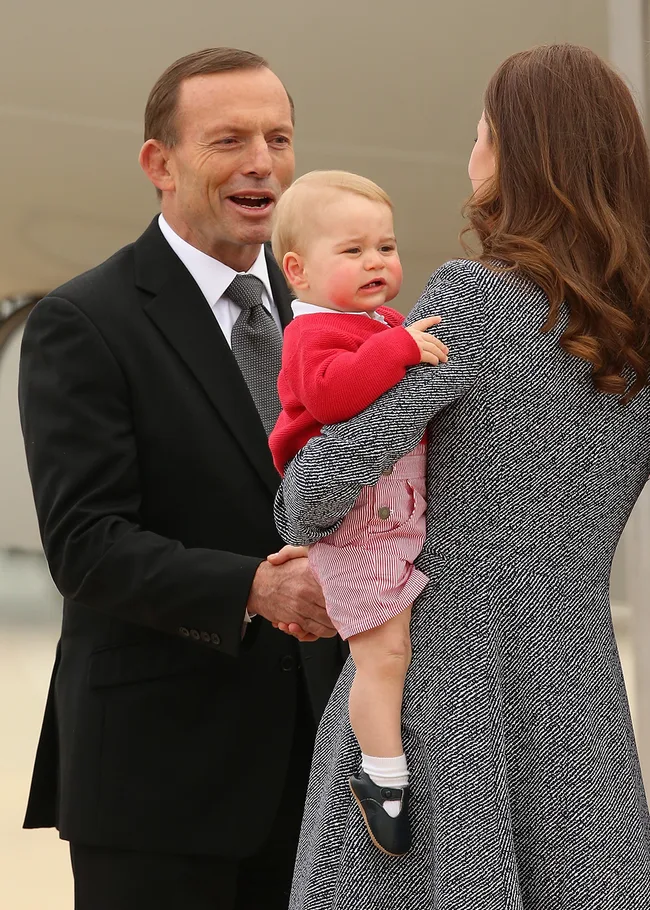 Man in suit interacts with woman holding a baby in a red sweater and striped shorts.