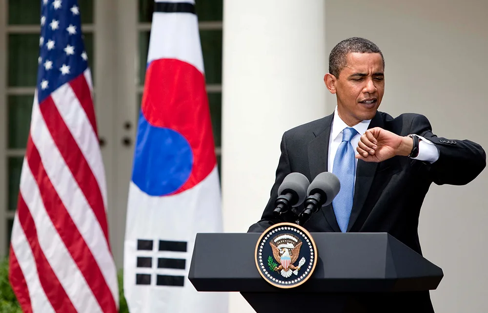 American man at podium checks watch between U.S. and South Korean flags.
