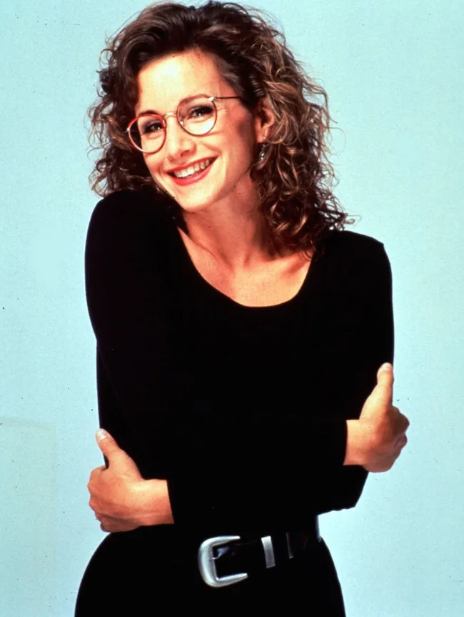 A woman with curly hair, wearing glasses and a black top, smiling with arms crossed.