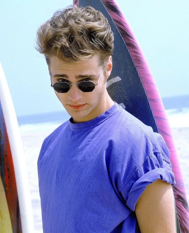 Person in a purple shirt and sunglasses standing by surfboards on a beach.
